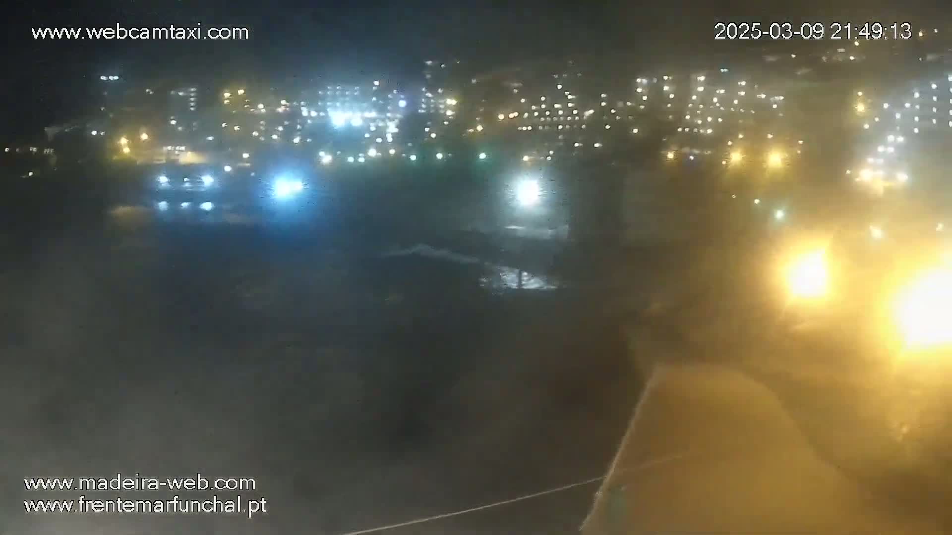 A nighttime, hazy view of a city waterfront showing lights from buildings and vehicles, with waves visible in the water.
