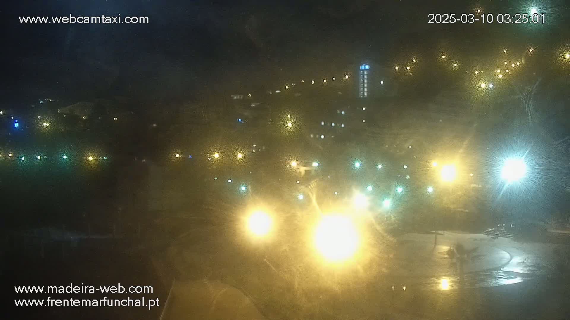 A nighttime view of a town illuminated by numerous lights, with a wet ground suggesting rain or recent precipitation.