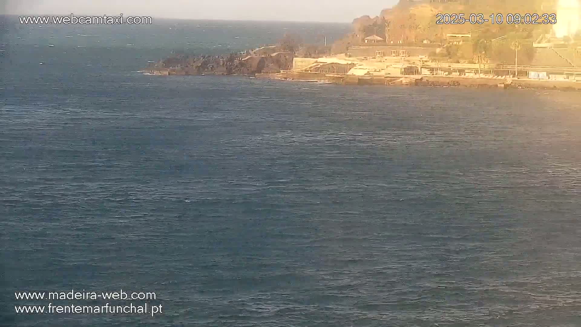 A calm ocean with a rocky shoreline featuring buildings and a small beach under a partly sunny sky.