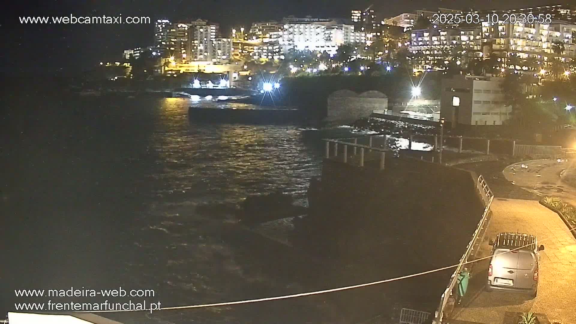 A nighttime view of a rocky coast with a van parked nearby, overlooking a dark body of water with moderate waves and illuminated buildings in the background.