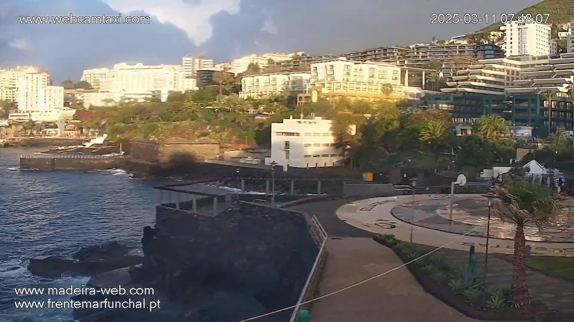 A coastal scene with a partly cloudy sky shows a collection of buildings overlooking a dark-colored, rocky shoreline and ocean.