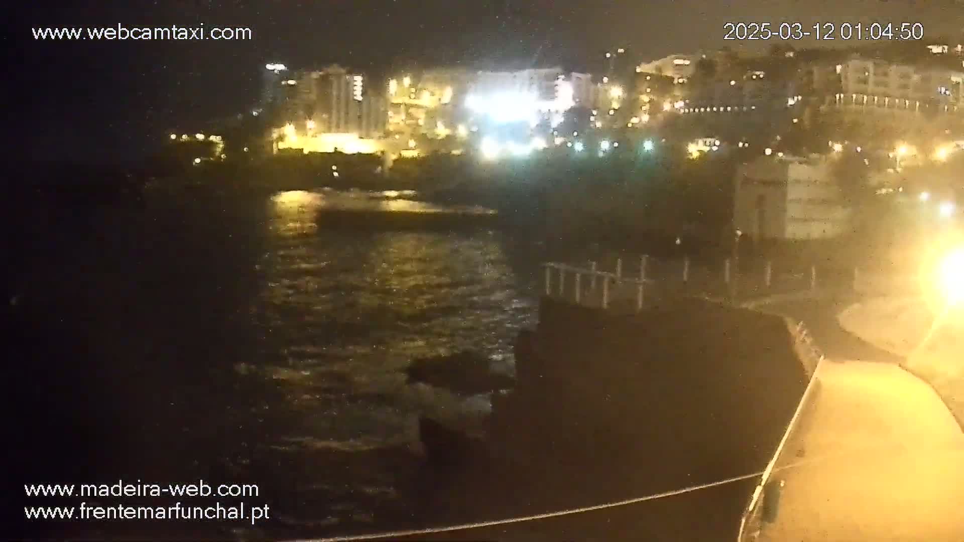 A nighttime view of a dark body of water reflecting the lights of buildings on a shore under a slightly overcast sky.