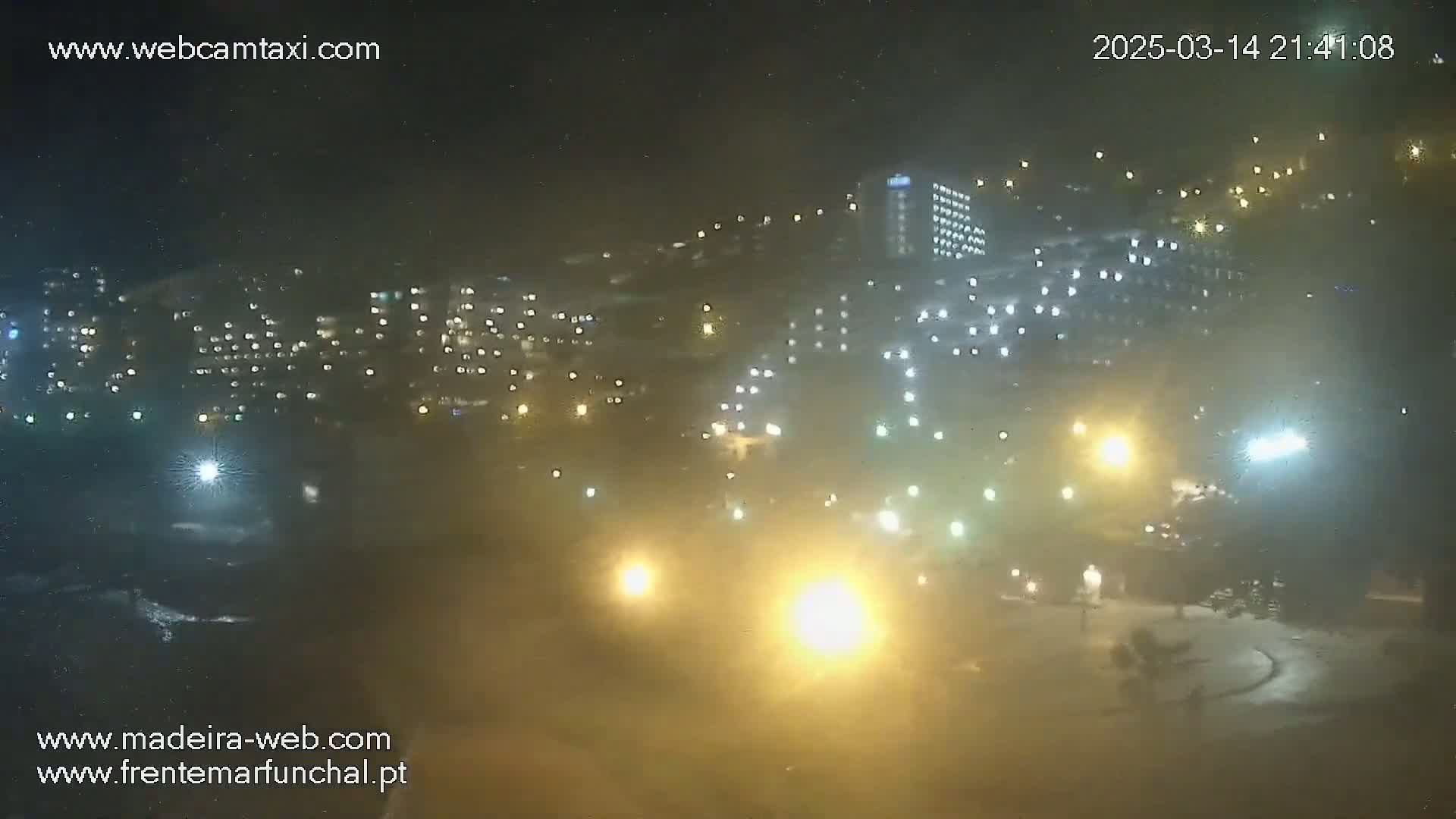 A hazy, nighttime view of a city illuminated by numerous lights, with buildings and structures on a hillside, under a dark sky.
