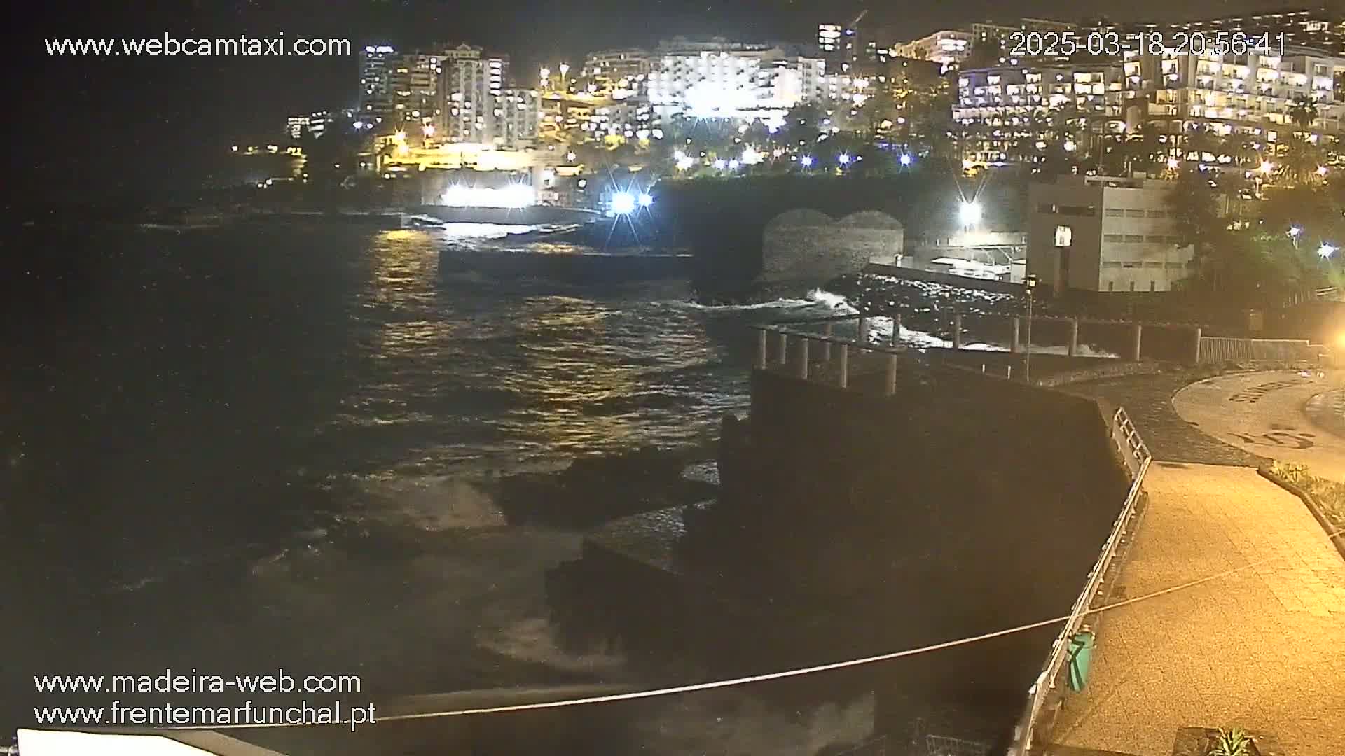 A nighttime coastal scene shows moderately rough water lapping against a rocky shore near buildings with lights on.