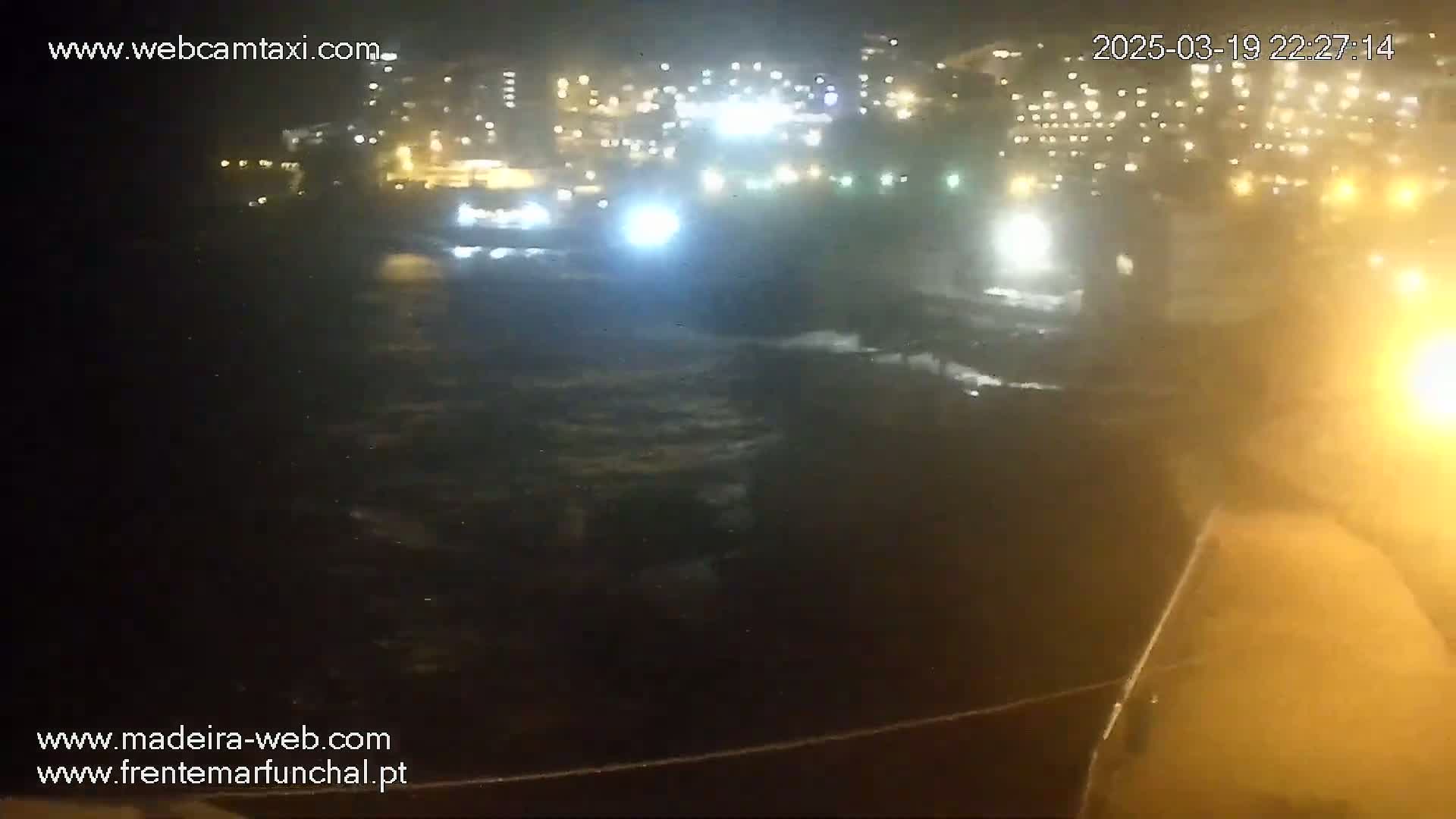 A nighttime view of a dark, choppy ocean with a brightly lit city in the background.