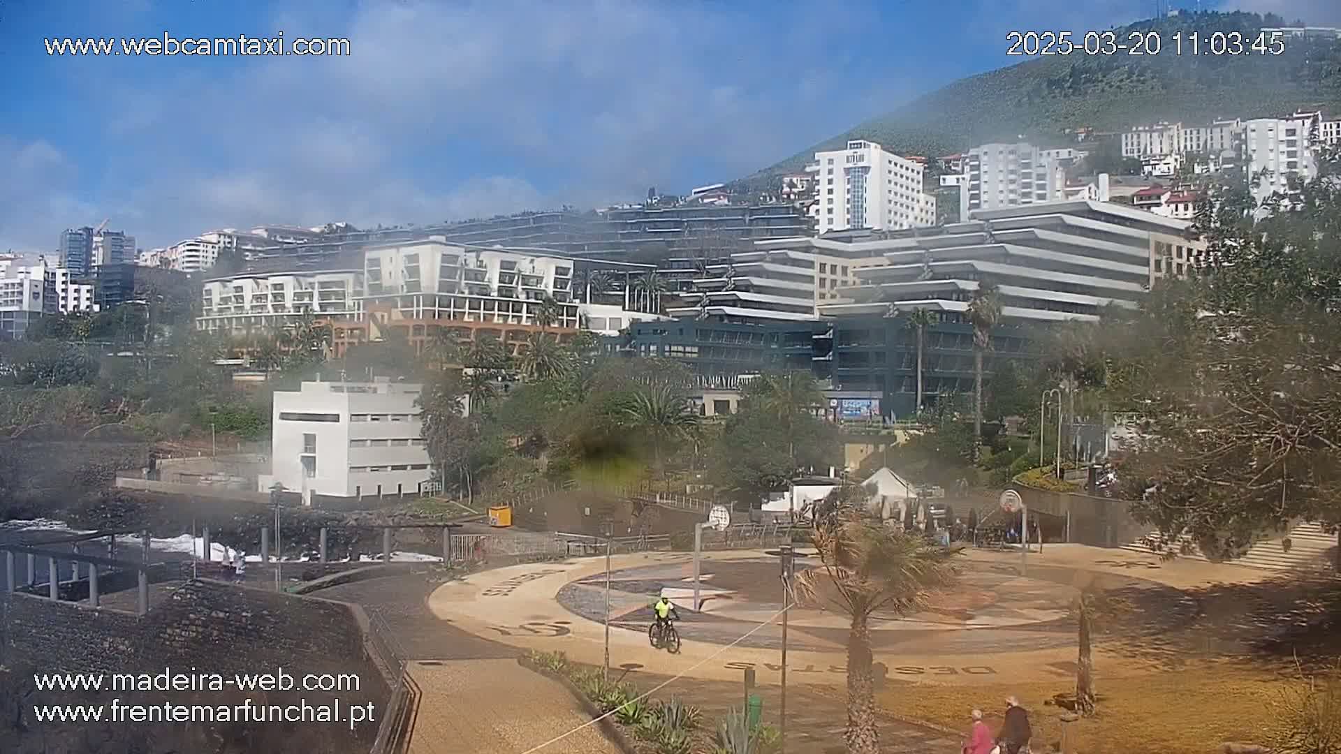 A hazy coastal cityscape with numerous buildings and a paved area featuring a compass rose design, a basketball court, and a cyclist, under a partly cloudy sky.