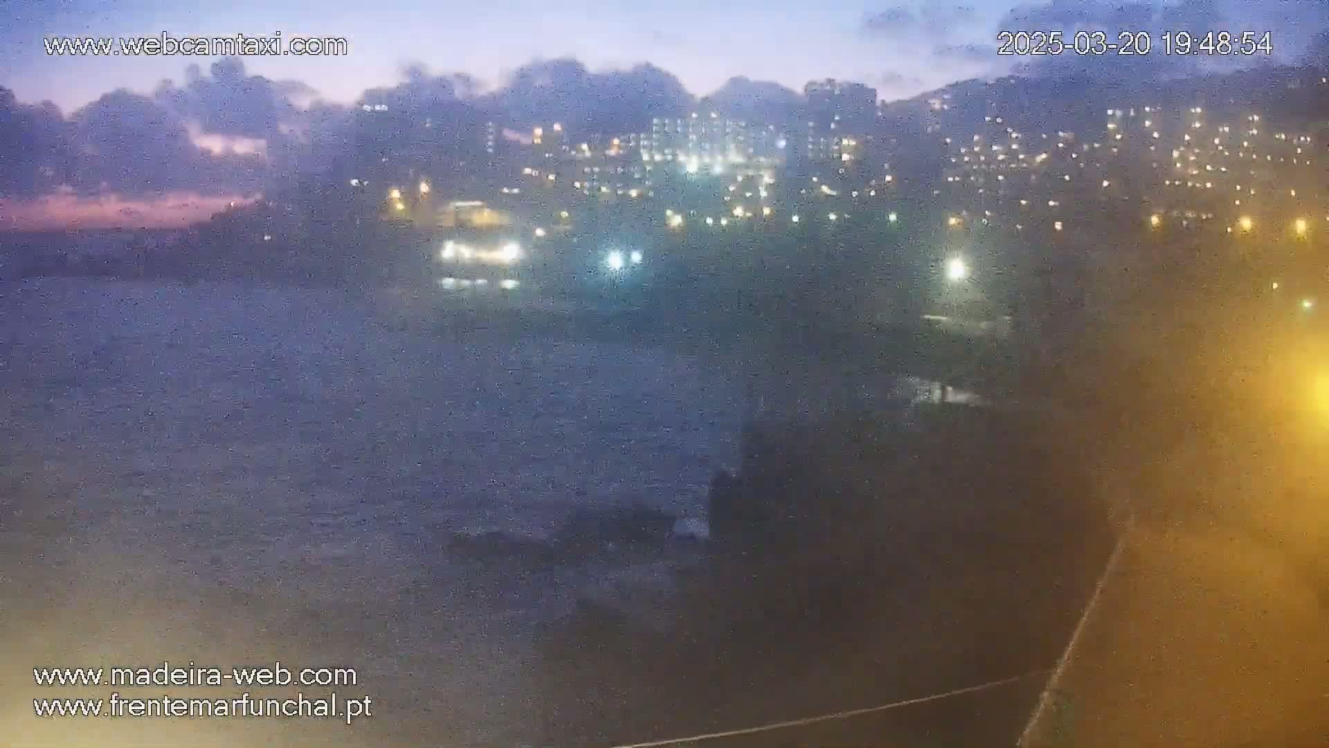 A nighttime view shows a dark, choppy ocean in front of a city with illuminated buildings under a cloudy sky.