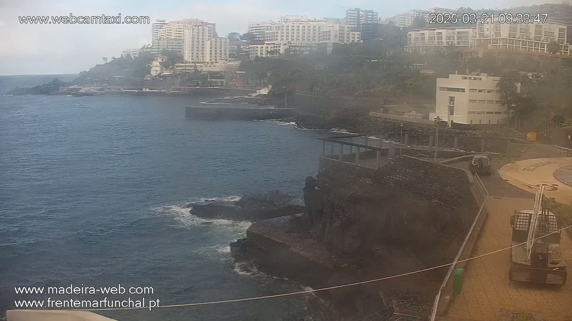 A hazy, overcast day reveals a coastal scene with dark-colored buildings, a rocky shoreline, and a calm ocean with slight wave action.