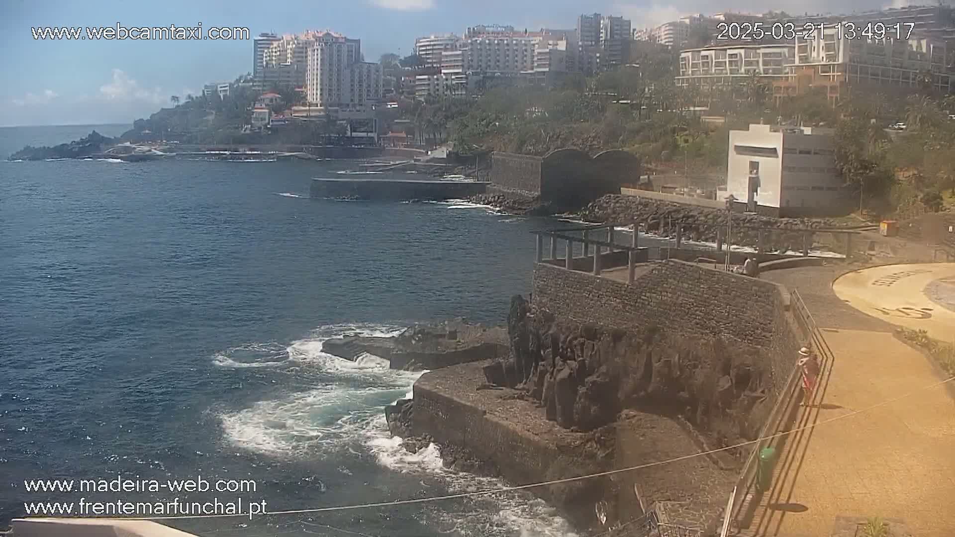 A rocky coastline with a calm ocean, some buildings in the background, and a few people walking along a paved walkway under a partly sunny sky.