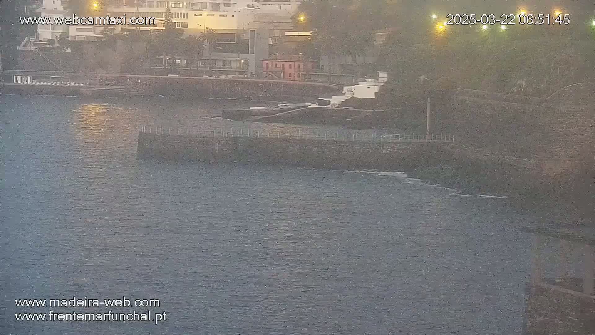 A calm body of water laps against a low stone wall near a waterfront building under a dusky sky.