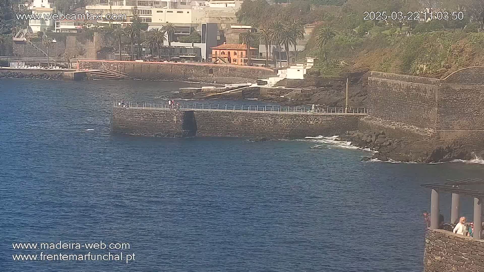 A stone wall extends into a calm blue ocean, with a few people walking on top and a small group of people standing on a stone structure to the right.
