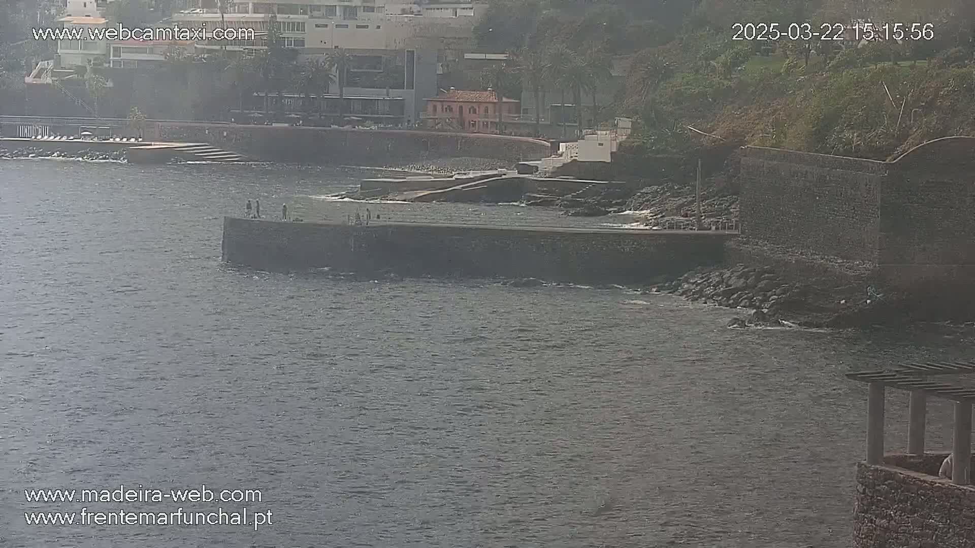 A hazy view of a rocky coast with a small structure and several people standing on a wall overlooking calm water.