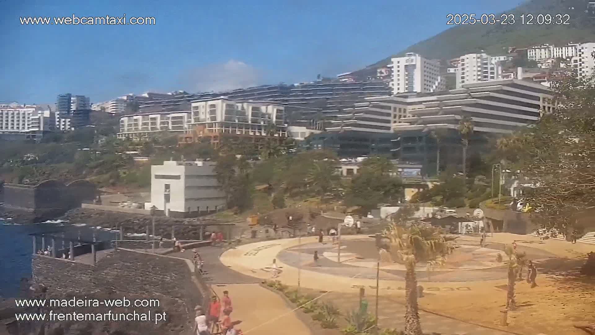 A sunny day overlooking a coastal city with multiple buildings, a paved plaza with people, and green vegetation.