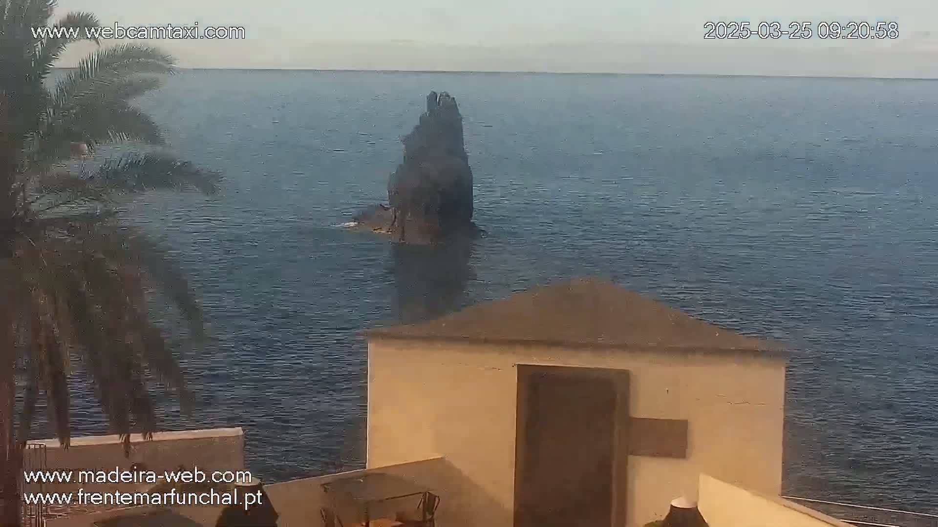 A dark, tall rock protrudes from a calm, blue ocean, viewed from a tan building with a small patio and partial palm tree in view.