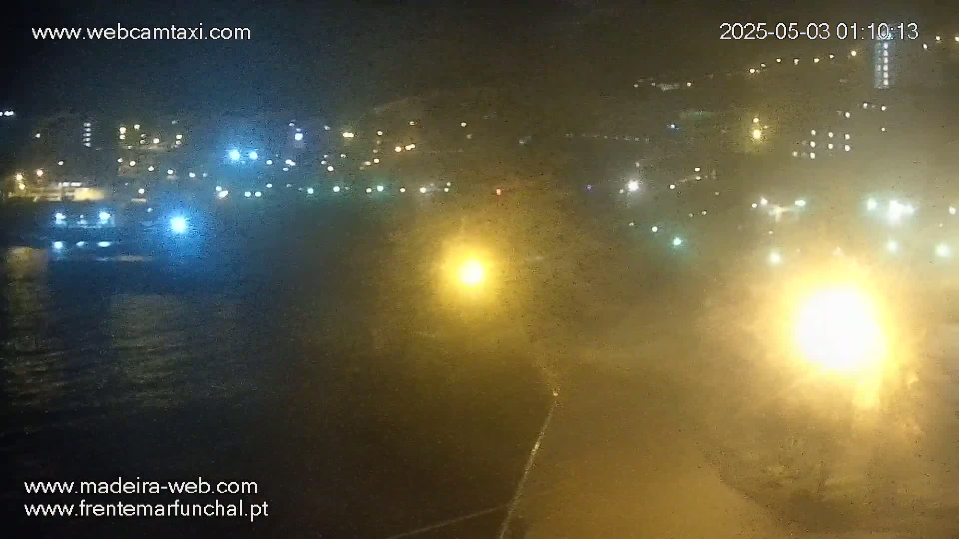 A hazy nighttime view of a waterfront city, illuminated by numerous lights, with a body of water in the foreground.
