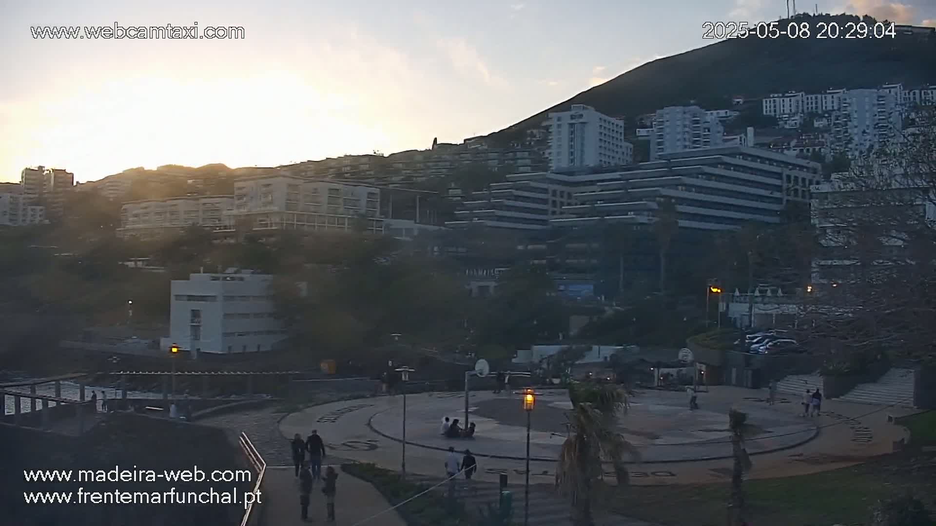 A twilight view of a city nestled against a hill, with people gathered in a paved plaza below a cluster of buildings.