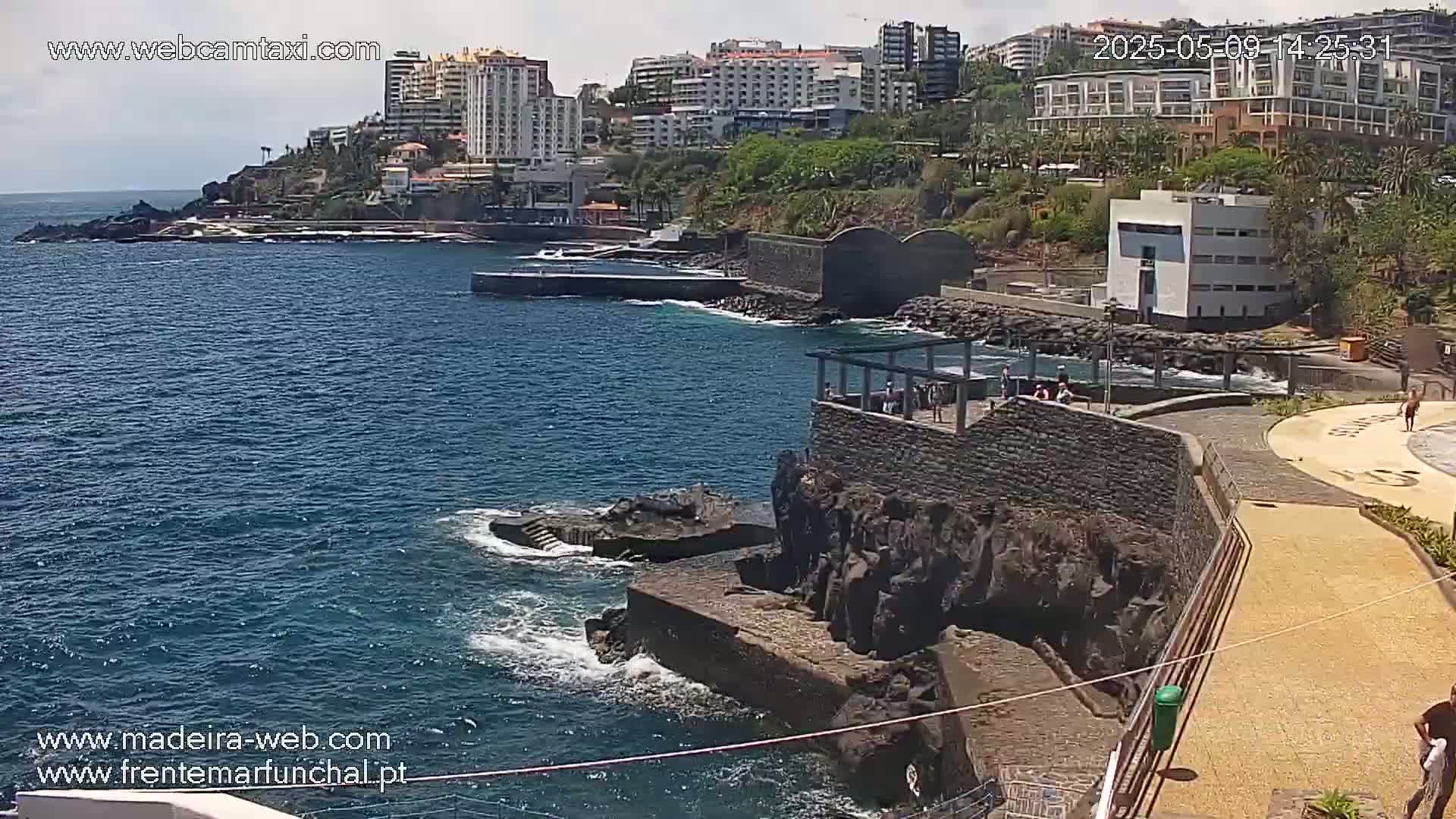 A coastal scene shows a rocky shoreline with a paved walkway, buildings in the background, and a calm ocean under a mostly sunny sky.