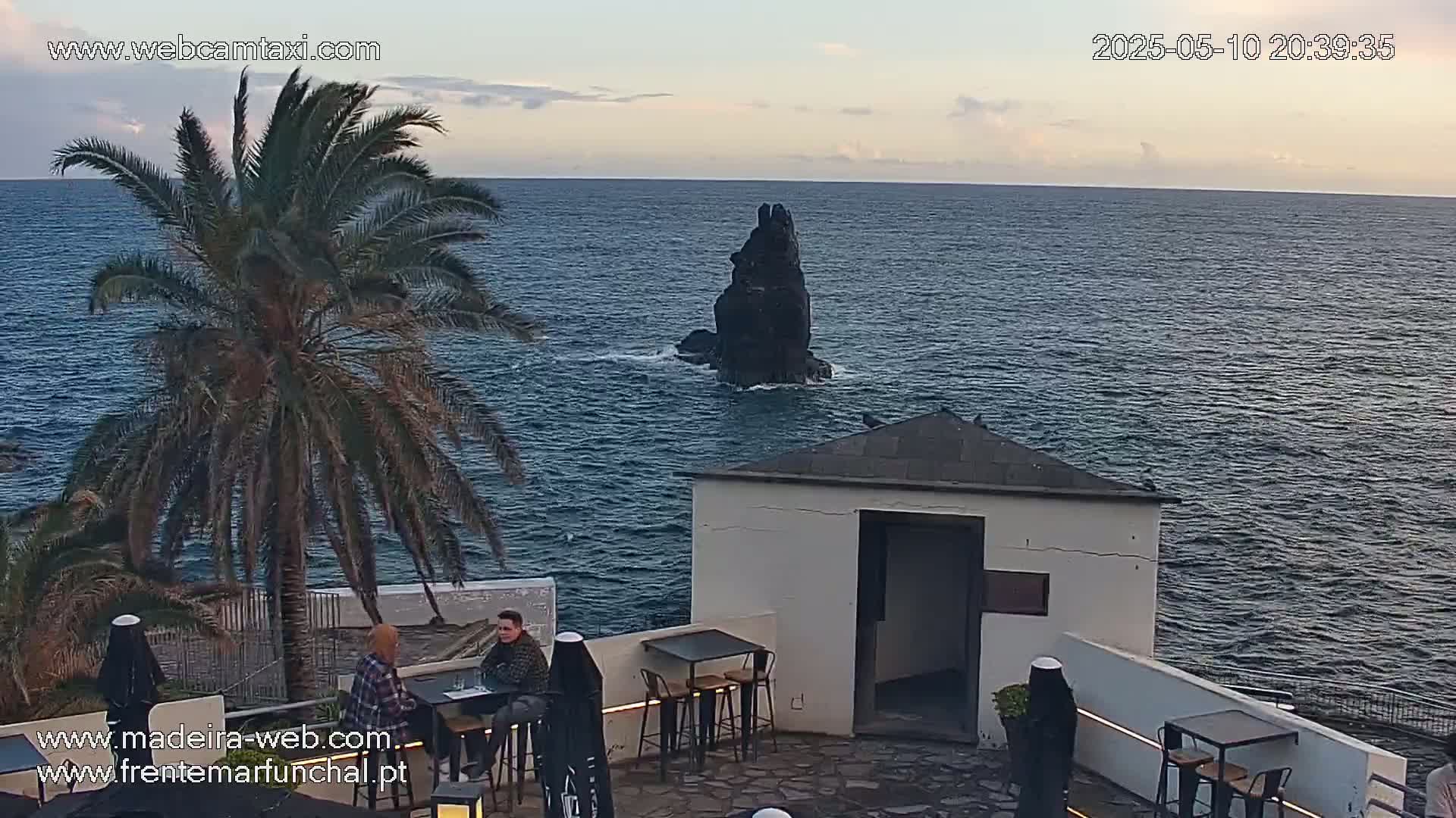 An outdoor patio with two people sitting at a table overlooks a calm ocean with a dark rock formation in the distance under a partly cloudy sky.