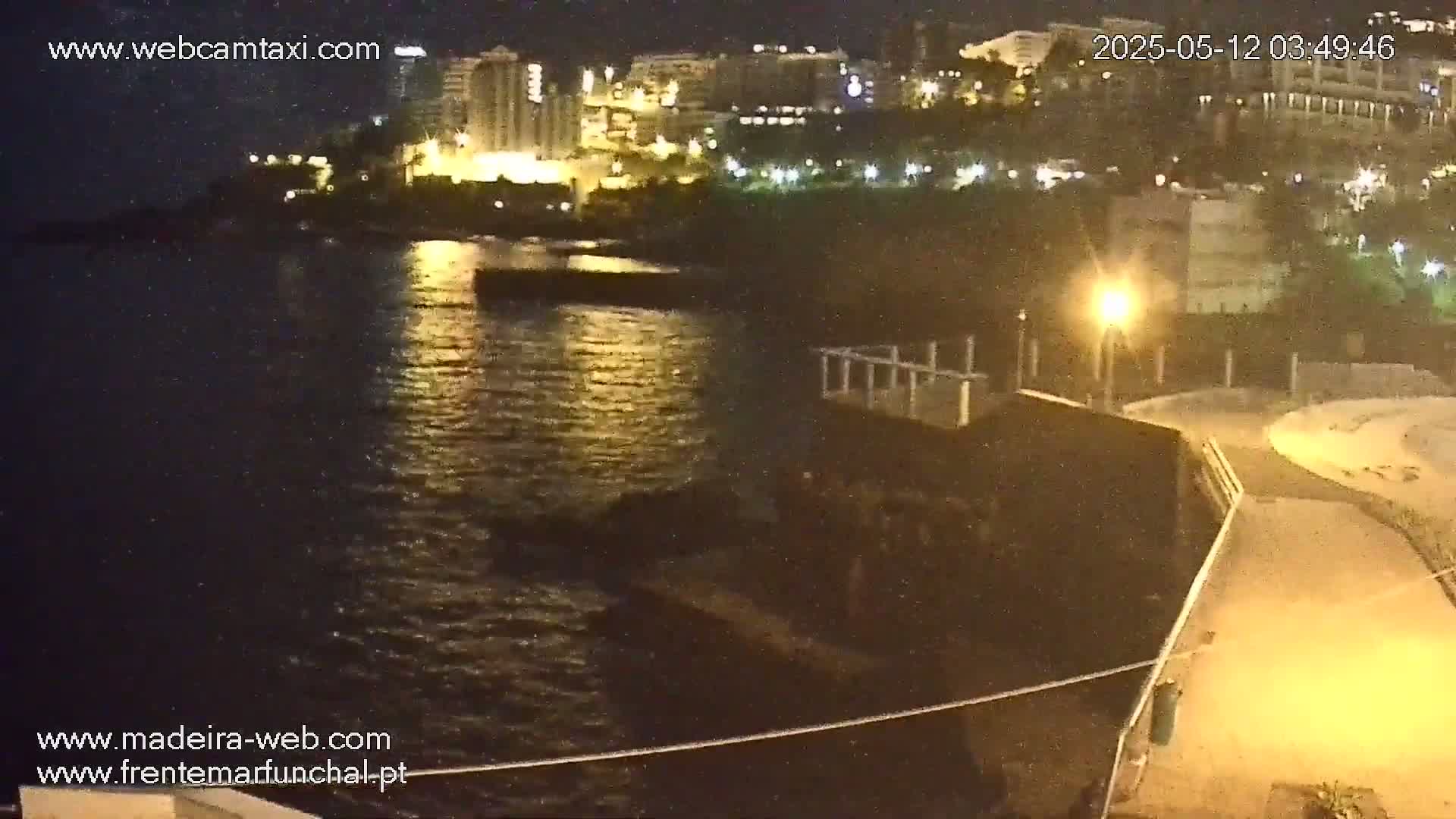 A nighttime view shows a dark body of water reflecting lights from buildings on the opposite shore, along with a paved walkway and a structure near the water's edge.