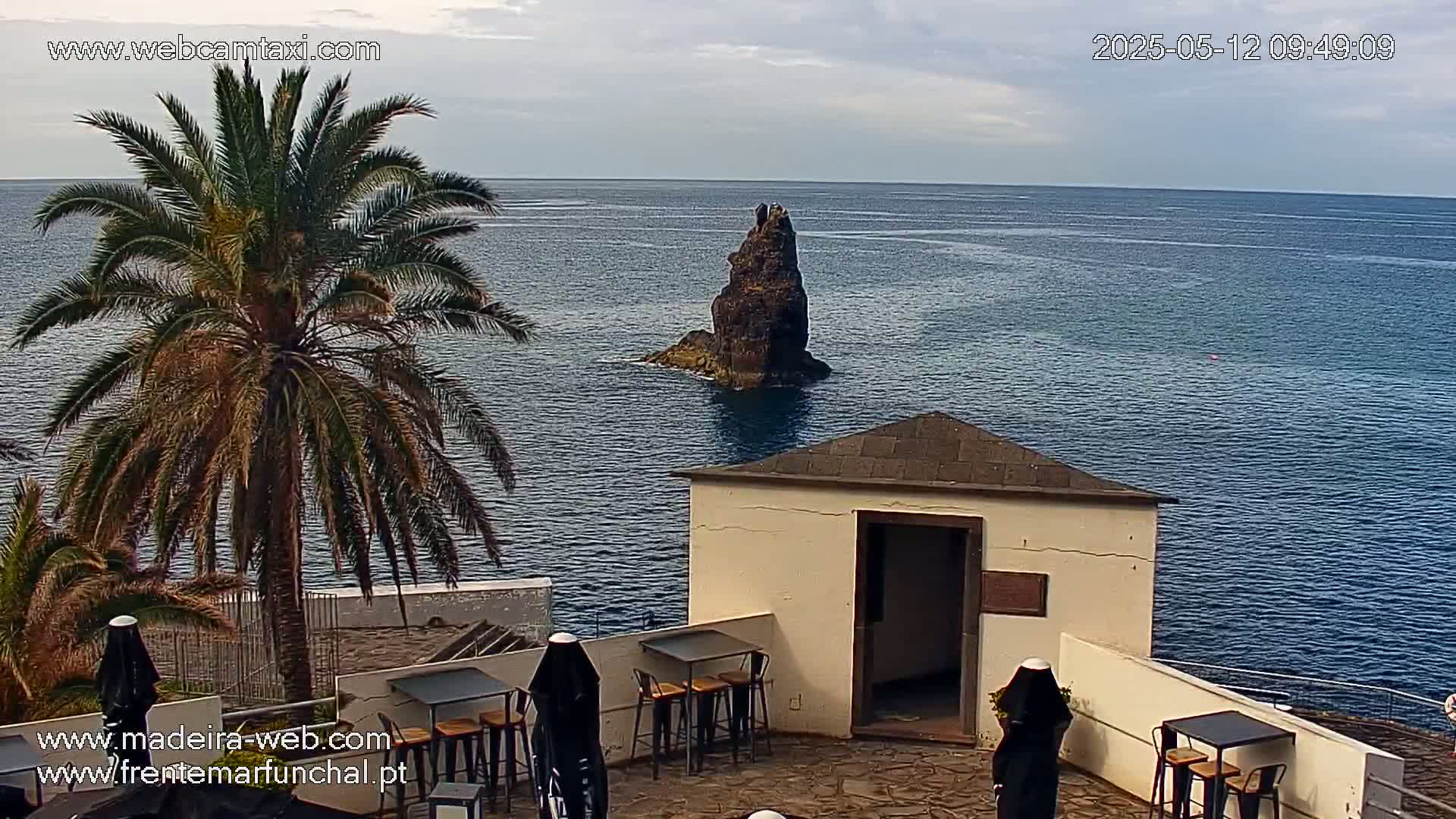 An outdoor patio with tables and chairs overlooks a calm ocean, where a lone rock formation sits in the water, under a partly cloudy sky.