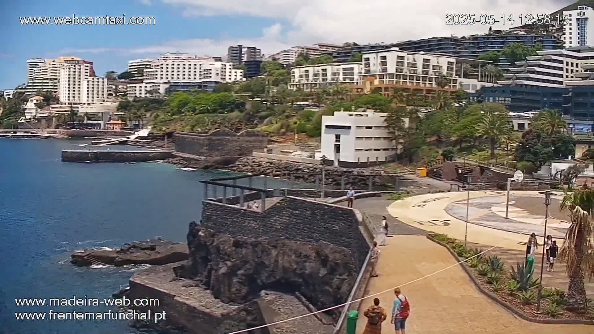 A seaside view of several hotels and buildings, a rocky coastline, a calm ocean, and people walking along a paved path on a sunny day.