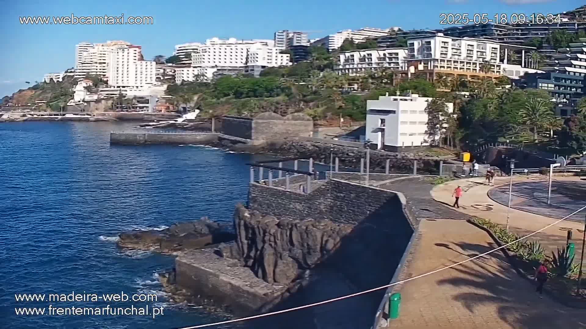 A coastal scene on a sunny day shows dark-colored stone structures near a body of blue water, with several buildings visible in the background.