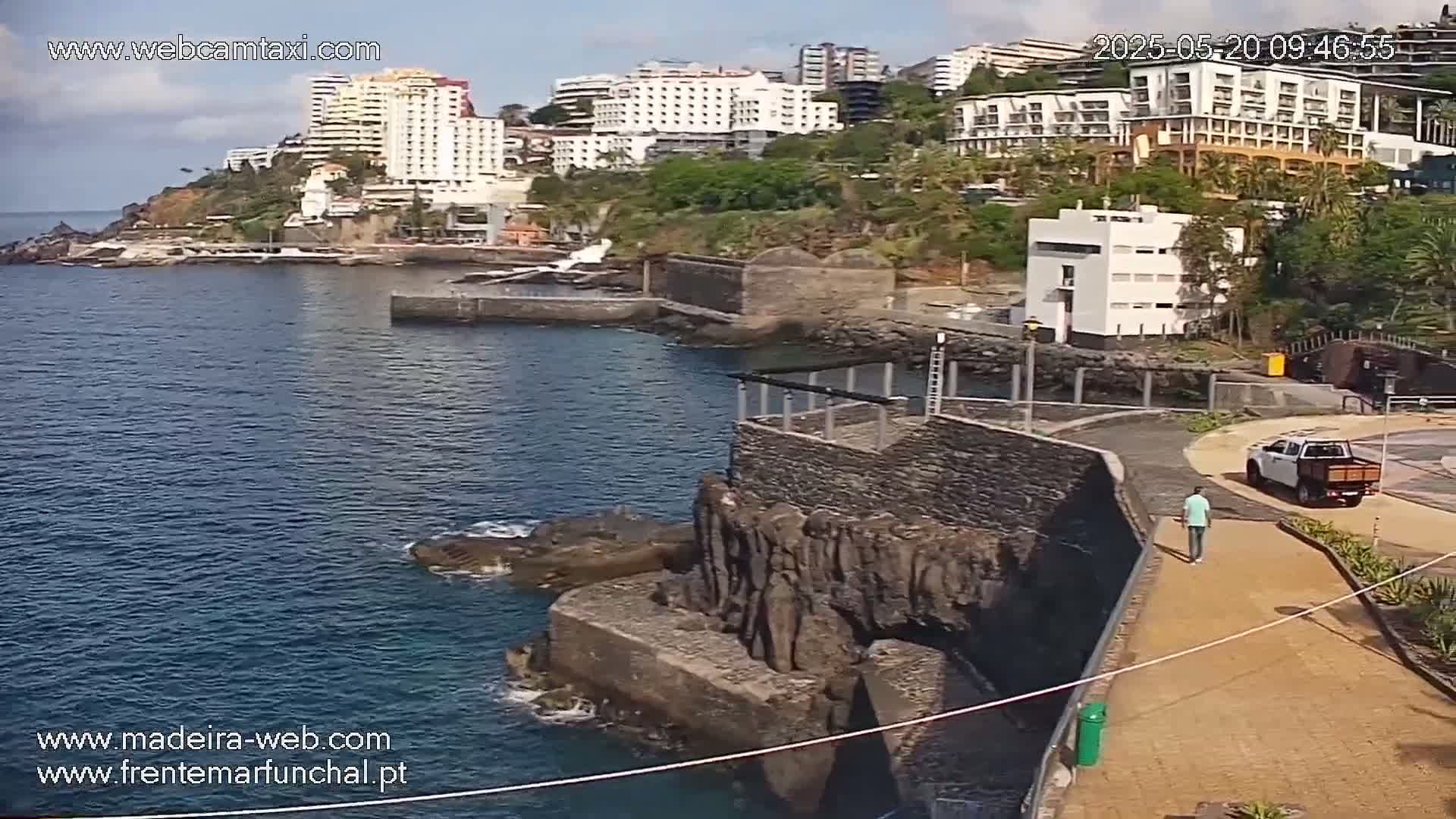 A man walks along a paved path overlooking a rocky coastline and calm blue ocean, with several buildings visible on a hillside in the background under sunny skies.