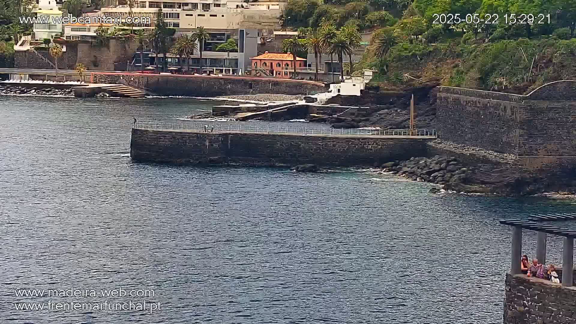 A stone pier extends into a calm body of dark water next to a rocky shoreline and buildings, with a few people visible on the pier and others seated in a stone structure.