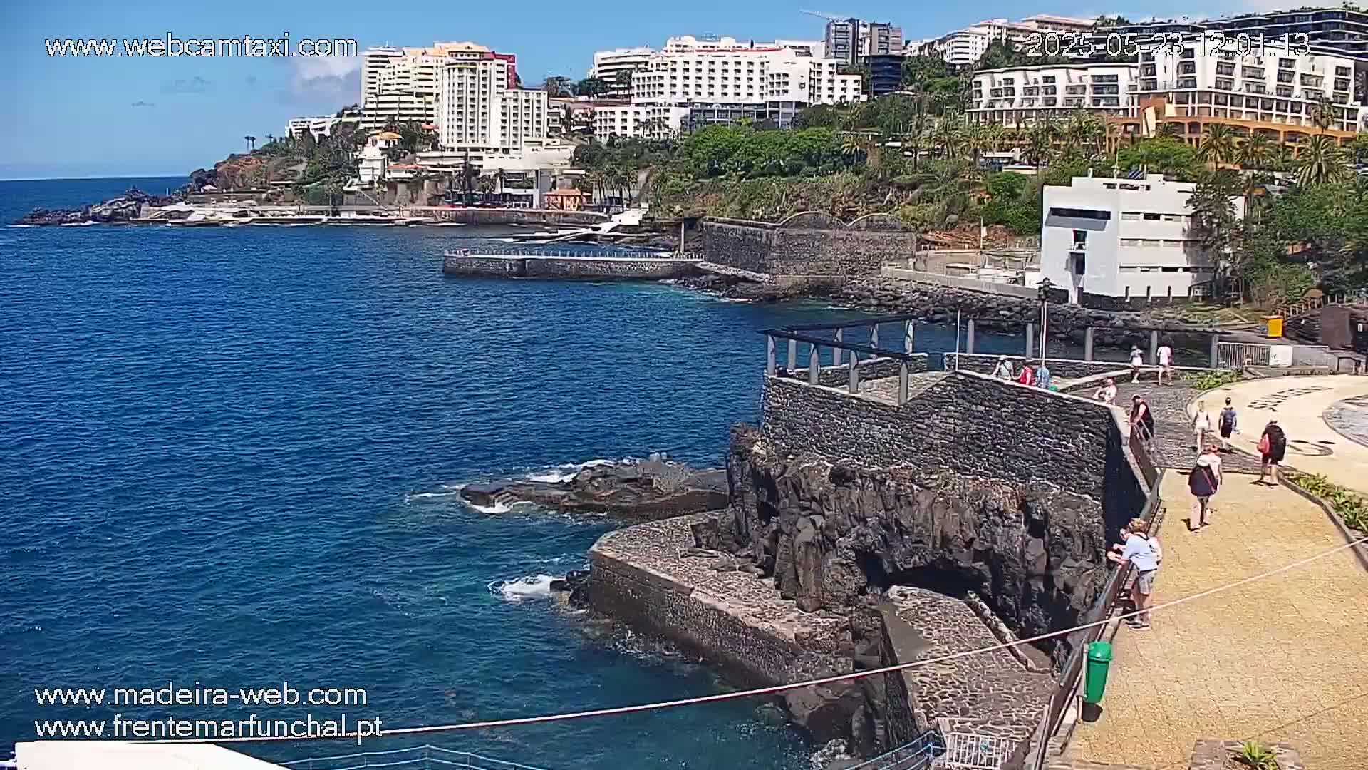 A rocky coast with a walkway and several people, overlooking a calm blue ocean and a cluster of hotels on a sunny day.