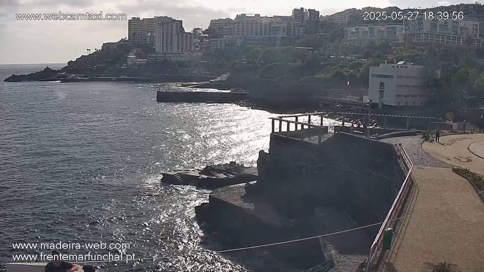 A rocky coastline with buildings in the background, under partly cloudy skies, shows a partly sunny day with glittering water.