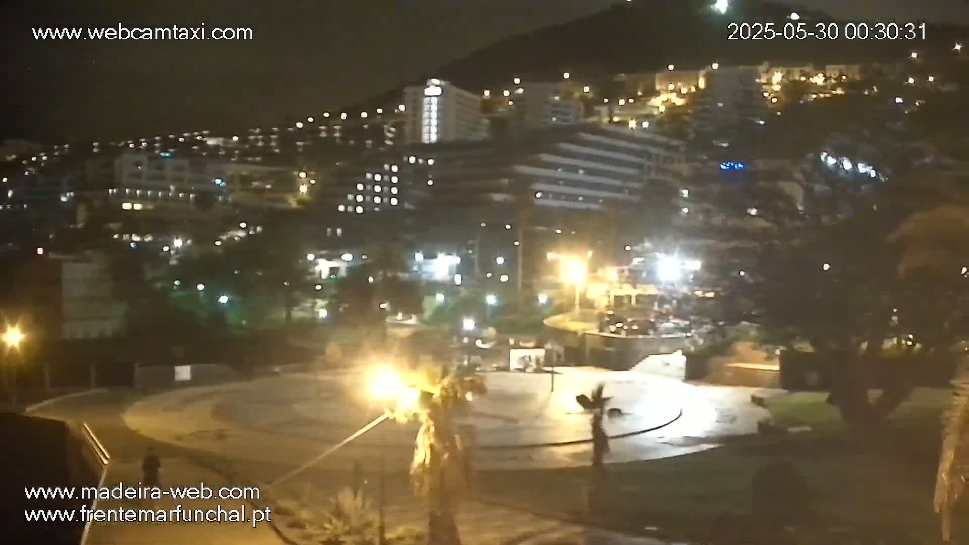 A nighttime view of a city with illuminated buildings and a plaza, under a clear sky.