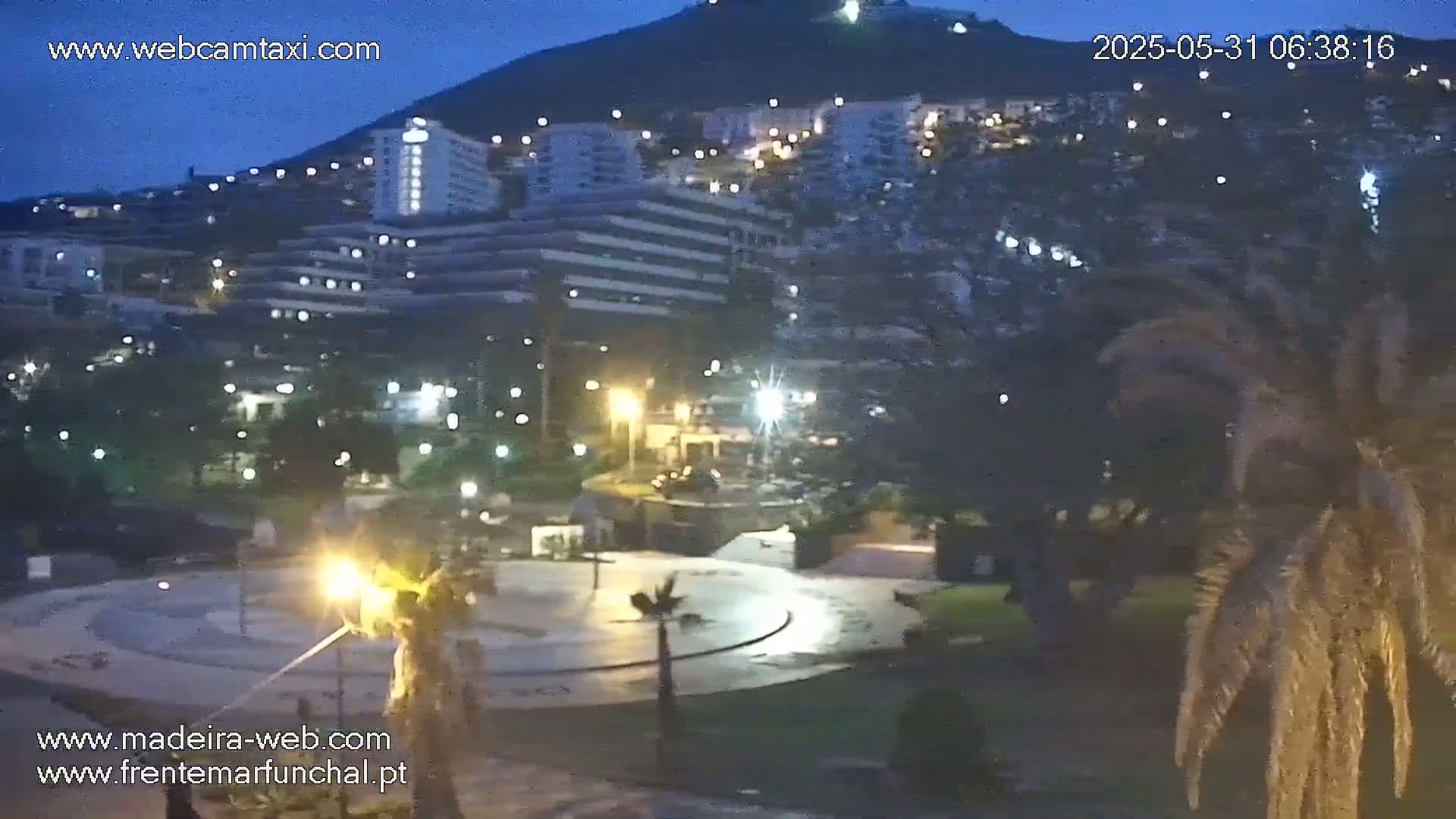 A nighttime view of a city with buildings illuminated against a dark mountain, under a twilight sky.