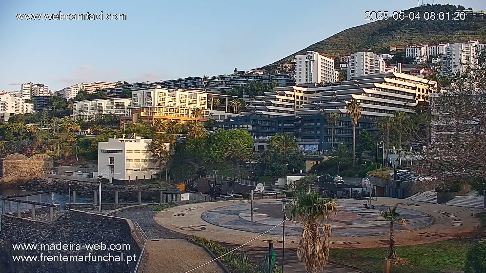 A hillside of multi-story buildings and hotels is seen at dawn under a clear sky, with a paved plaza and basketball court in the foreground.