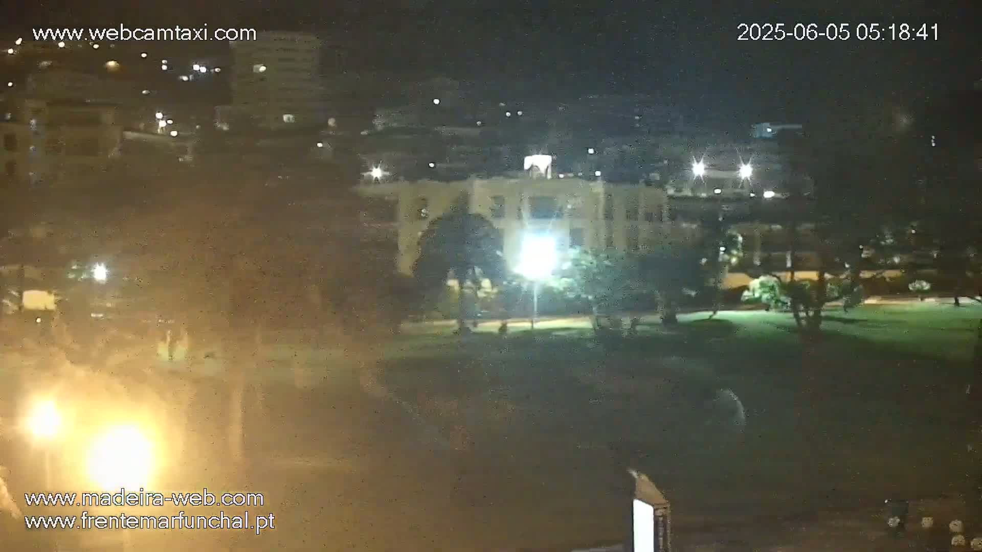 A nighttime view of a park with a building and streetlights in the background, under a slightly hazy sky.