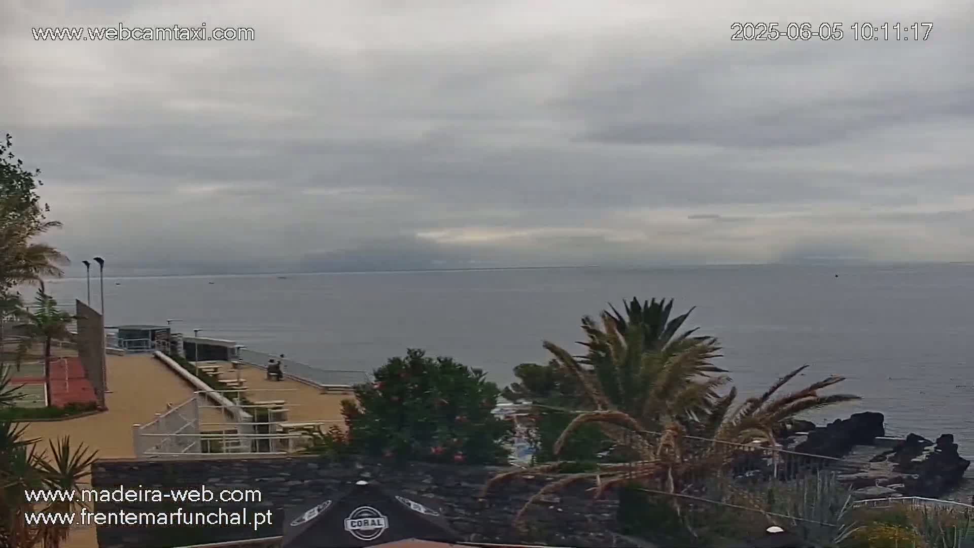 An overcast sky hangs over a calm ocean viewed from a coastal area with palm trees, a patio with seating, and a tennis court.