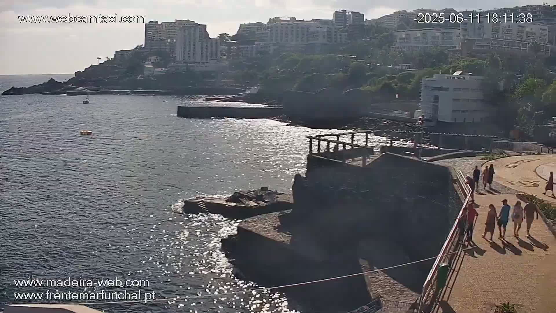 A rocky coastline with buildings in the background, a calm ocean, and several people walking along a paved path on a sunny day.
