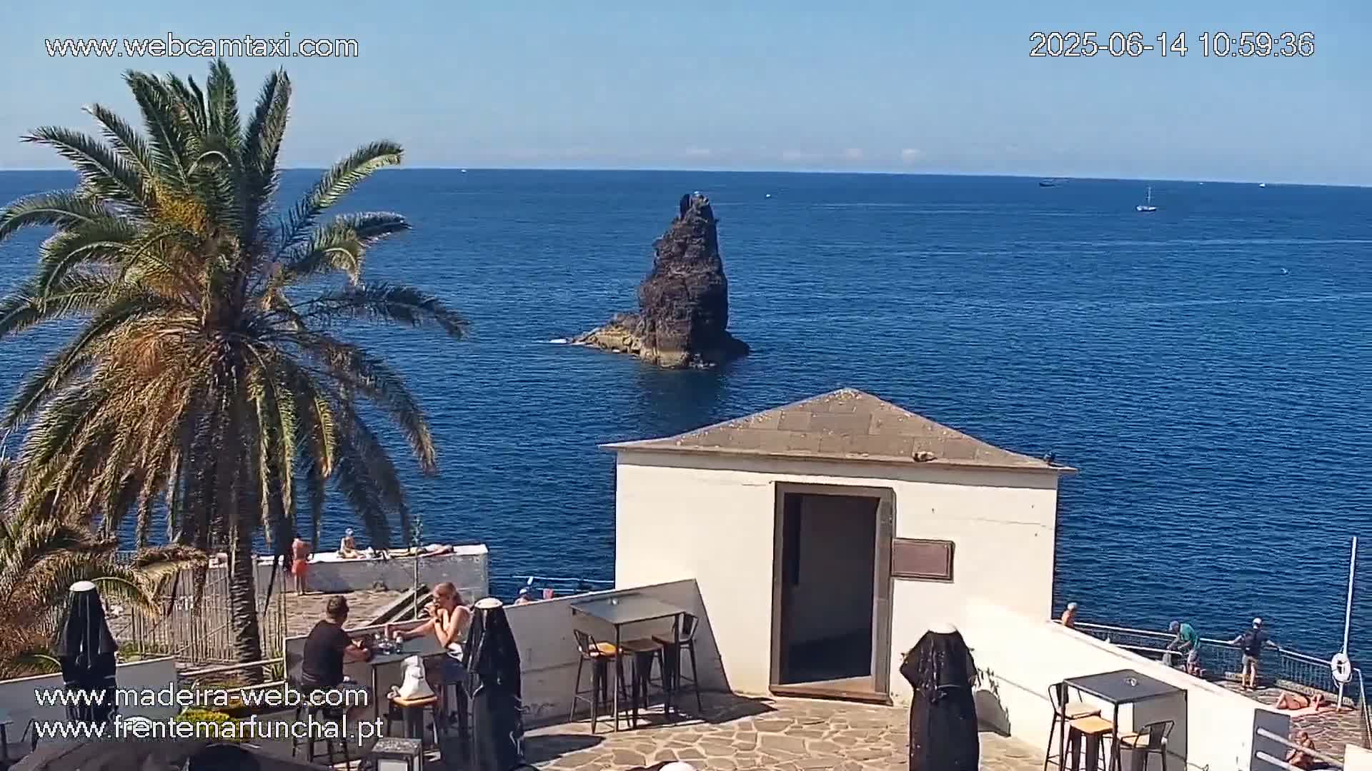 A seaside patio with outdoor seating overlooks a dark rock formation in a calm blue ocean under a clear sunny sky.