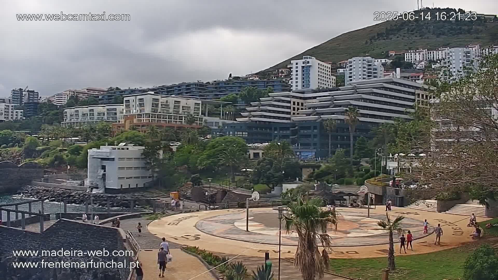 A coastal city scene under an overcast sky shows numerous multi-story buildings on a hillside, a paved plaza with people walking and playing, and the ocean partially visible in the foreground.