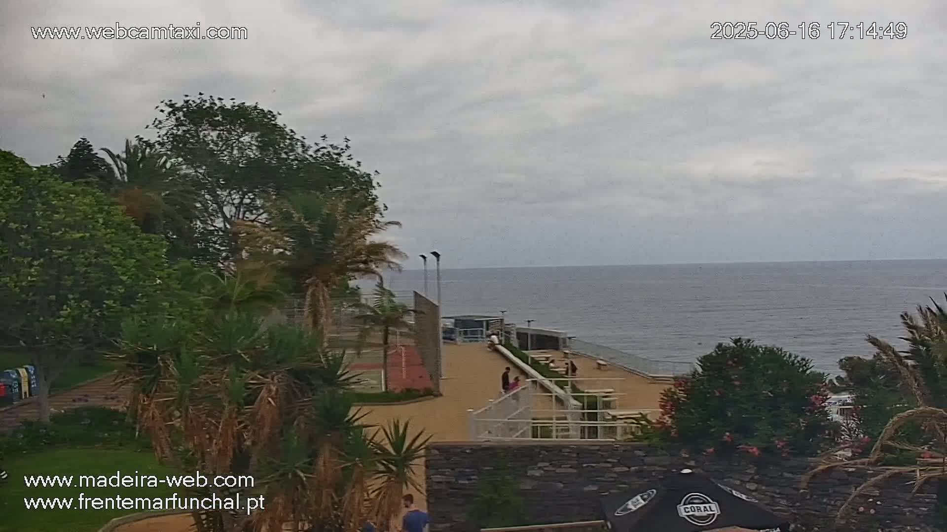 An outdoor scene shows a paved area with a small sports court overlooking a calm ocean under an overcast sky.