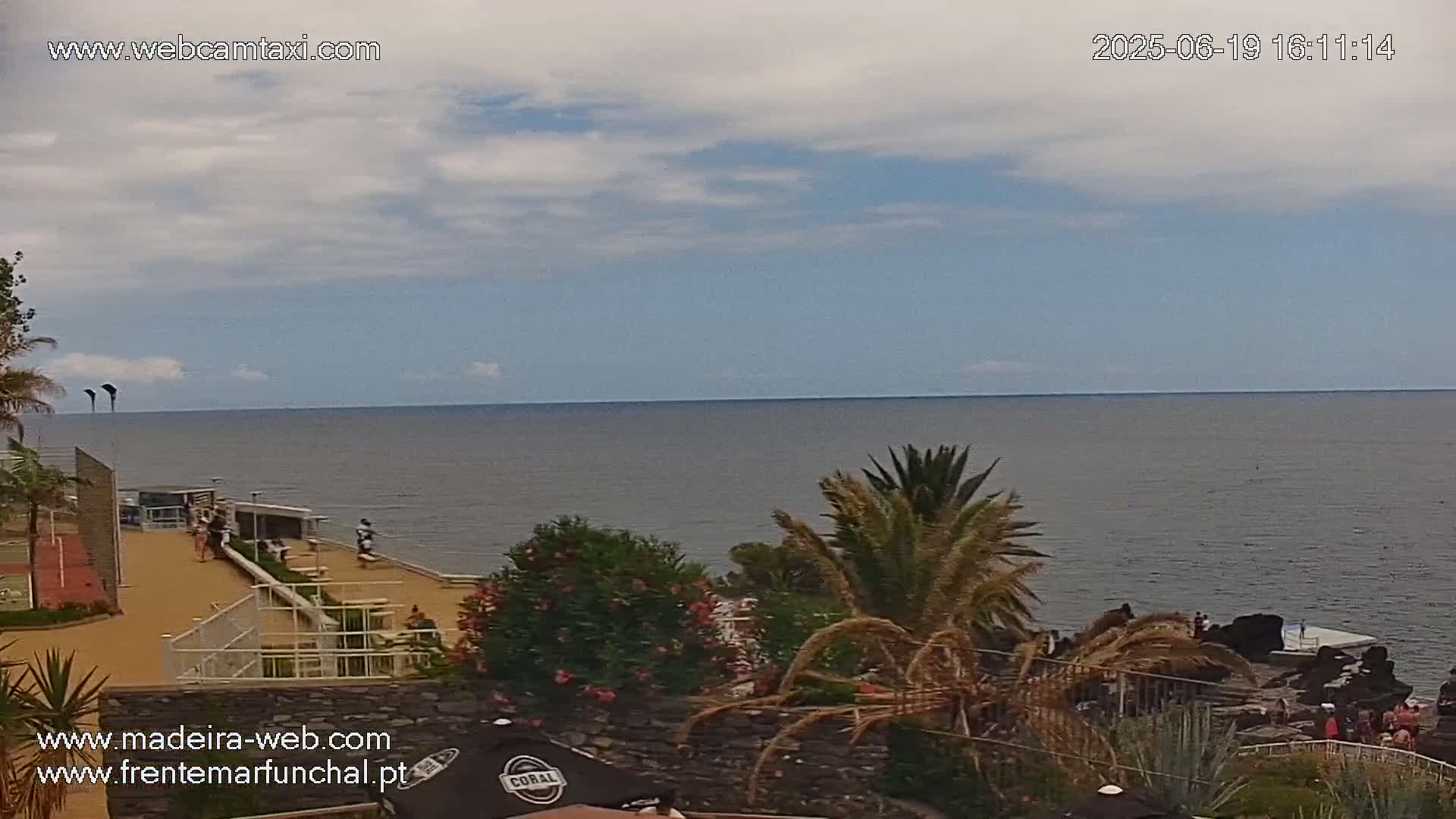 A partly cloudy day overlooking a calm ocean from a coastal viewpoint with vegetation and a few people near the water.
