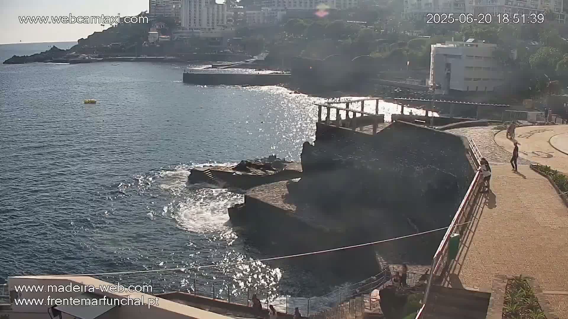 A rocky coastline with a partly sunny day and calm ocean waves, featuring a walkway with several people and buildings in the background.