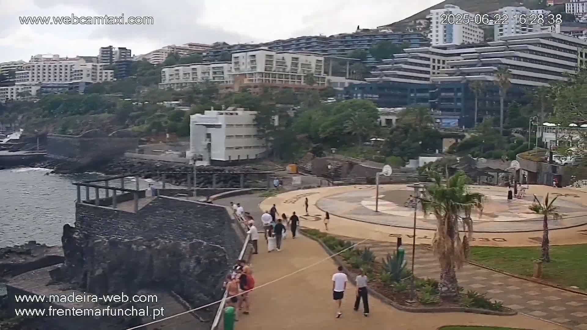 A coastal area with several multi-story buildings, a paved walkway with people, a basketball court, palm trees, and rocky shoreline under an overcast sky.