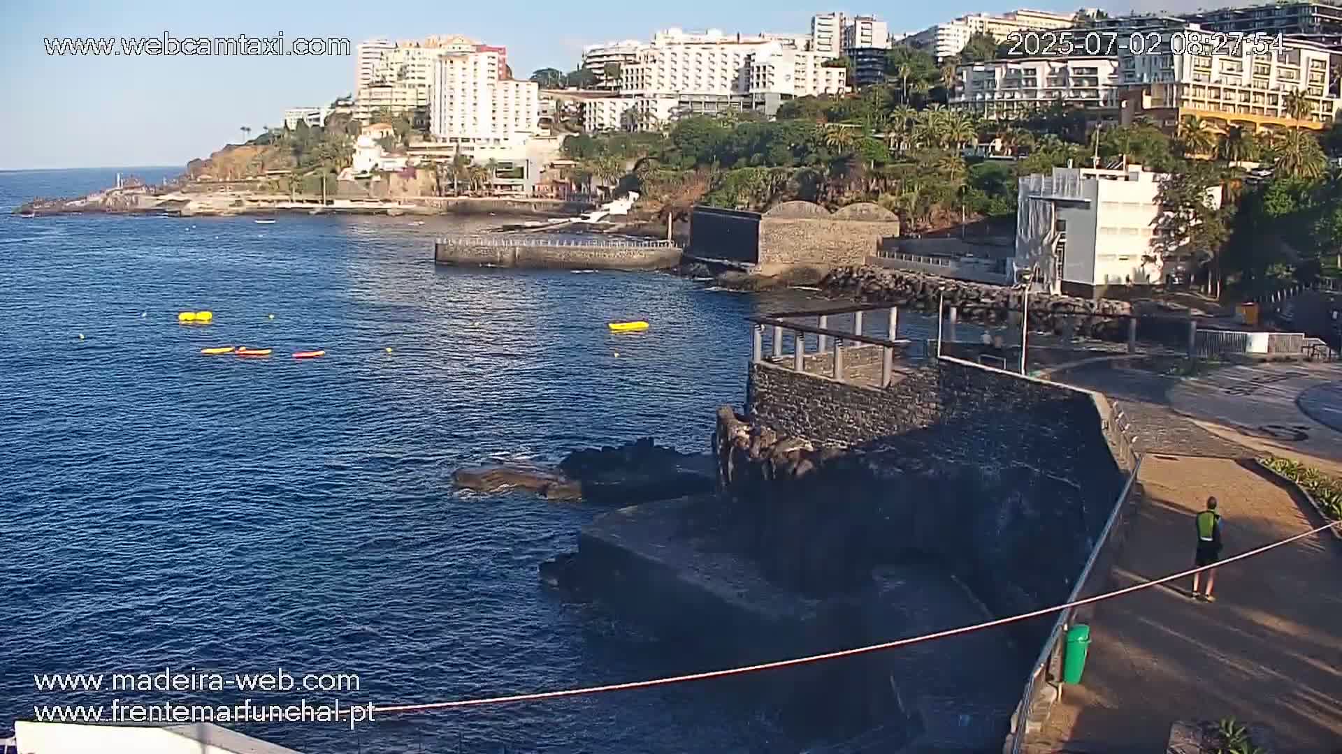 A sunny day at a rocky coastline shows several kayaks and paddleboards in the water near buildings and a paved walkway.