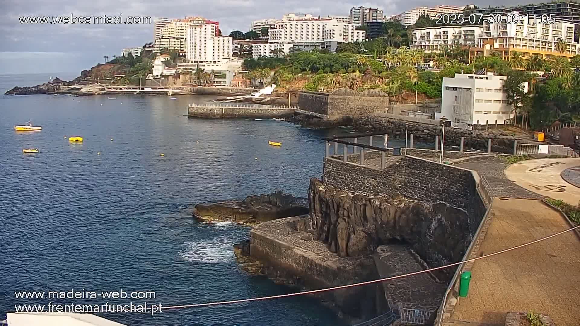 A mostly sunny coastal scene shows a calm body of water with several small boats, rocky jetties, and buildings in the background.