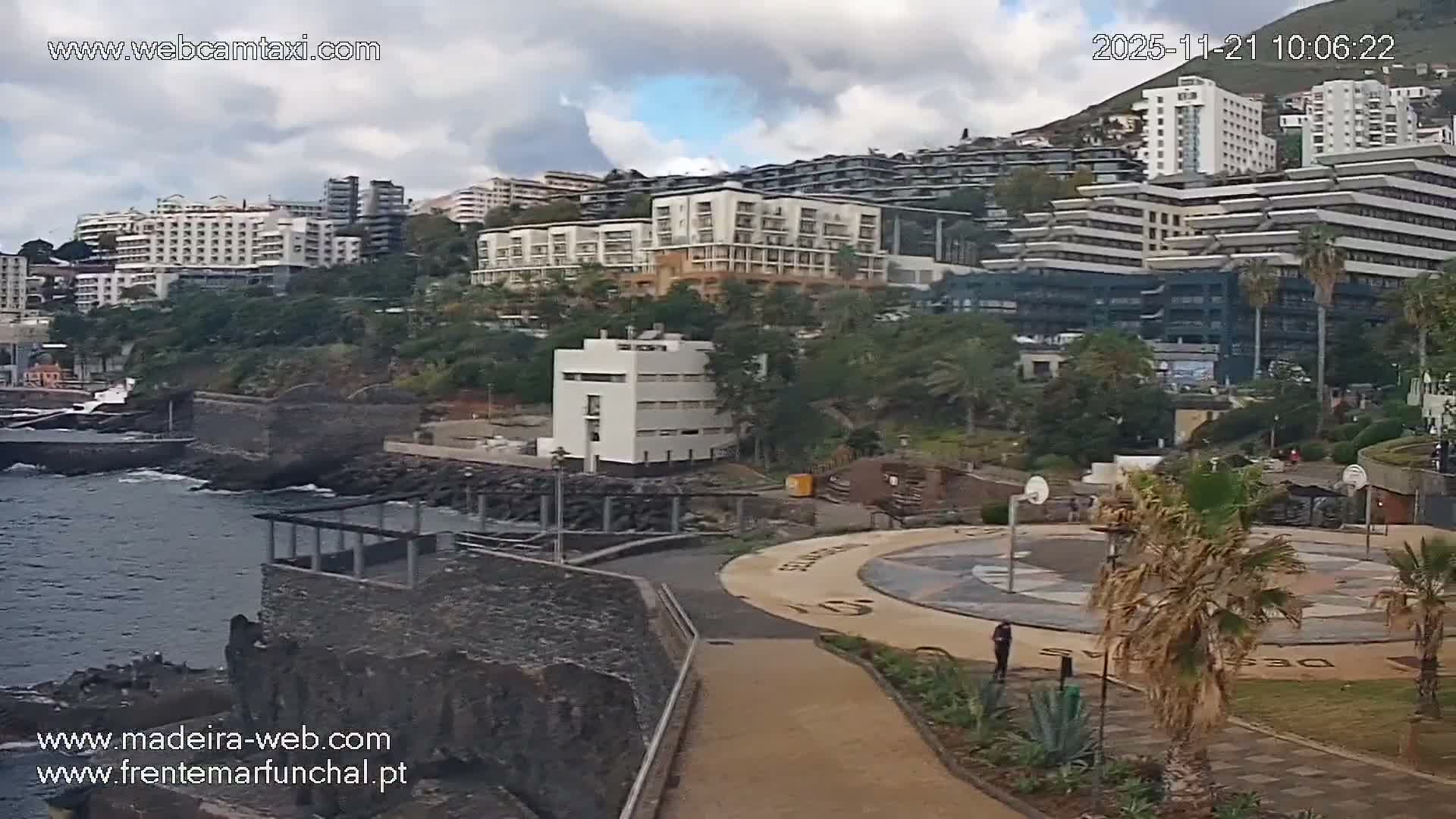 A coastal city scene unfolds under a partly cloudy sky, featuring a modern, multi-story building in the foreground with a paved promenade and palm trees, leading to an expansive view of numerous buildings cascading up a steep, vegetated hillside.