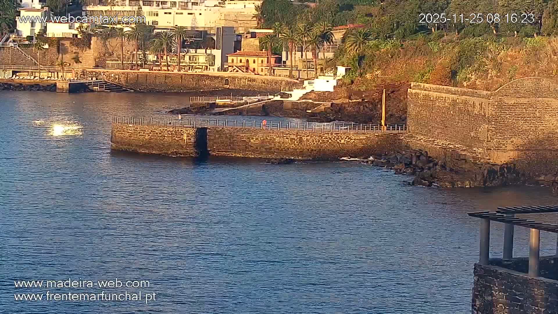 Under clear, sunny skies, a calm blue coastal inlet features stone seawalls and platforms, residential buildings and lush palm trees along a steep, rocky shoreline, all bathed in warm golden light.