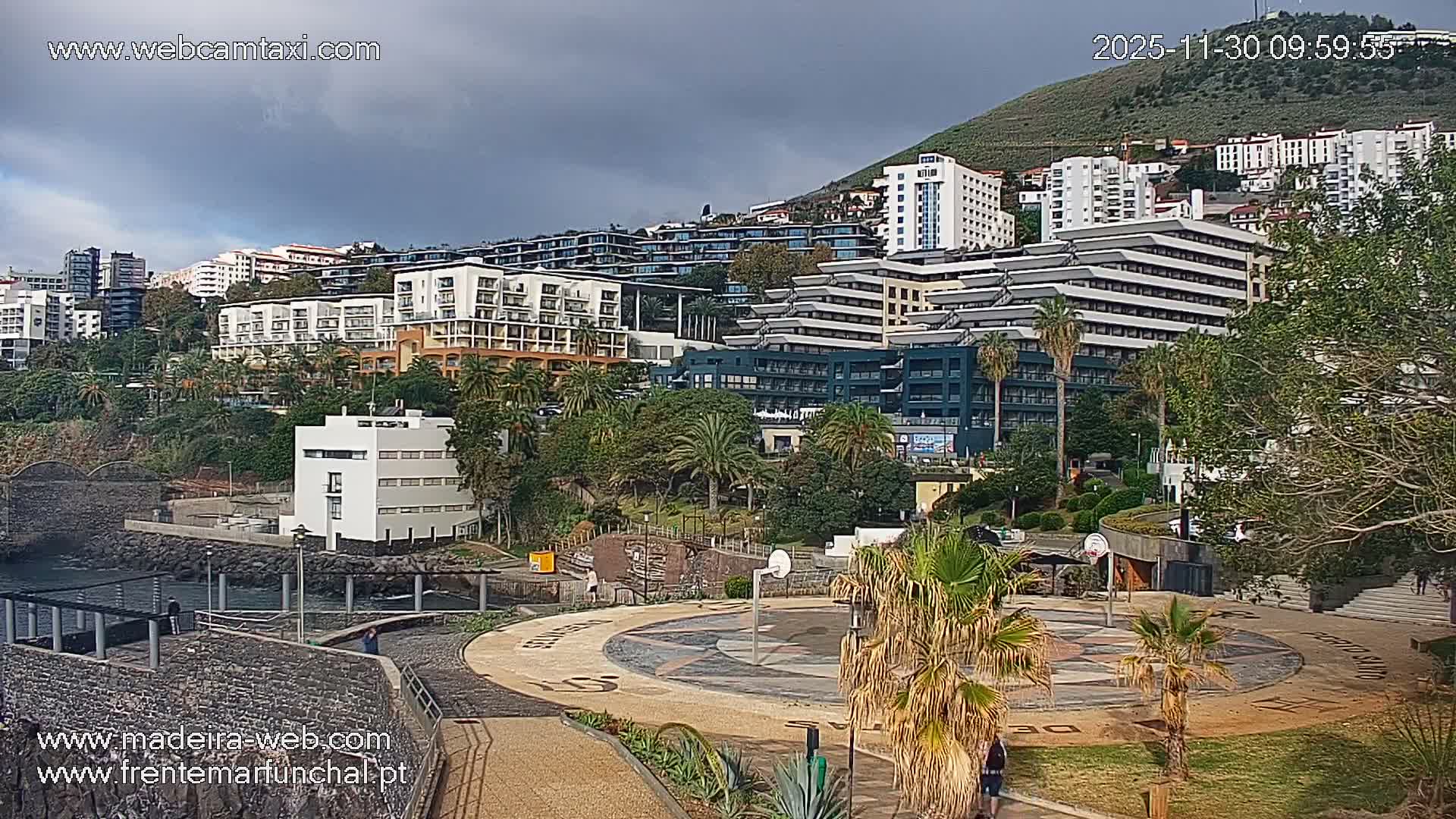 A modern coastal city, built into a lush green hillside dotted with multi-story buildings and palm trees, overlooks a dark, calm body of water with a white building on its shore and a patterned circular plaza in the foreground, all under an overcast sky.