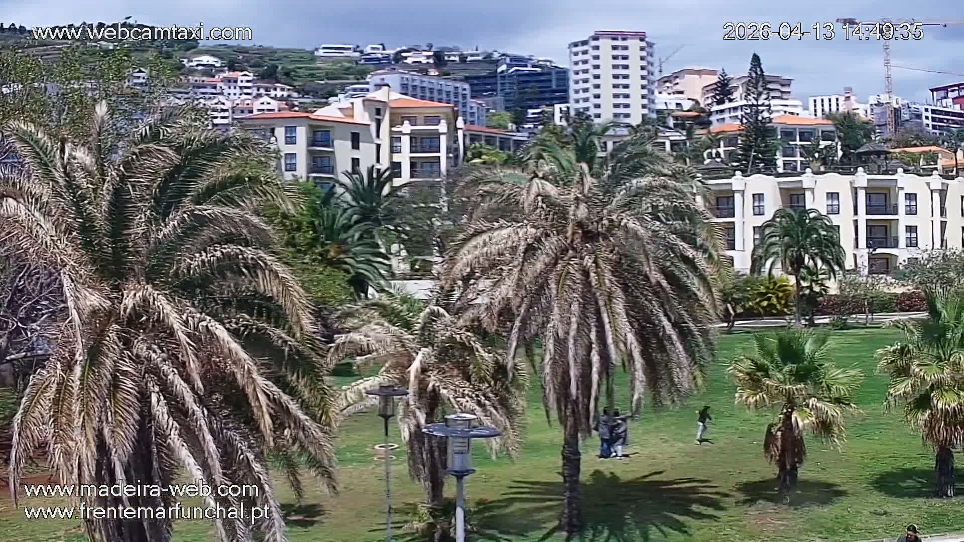A modern coastal city, built into a lush green hillside dotted with multi-story buildings and palm trees, overlooks a dark, calm body of water with a white building on its shore and a patterned circular plaza in the foreground, all under an overcast sky.