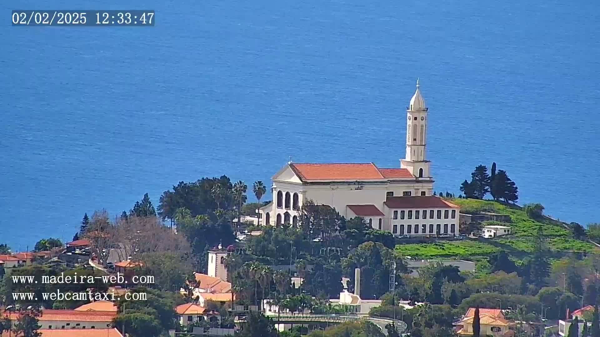 A white church with a tall bell tower sits on a small hill overlooking a calm blue ocean under a sunny sky.