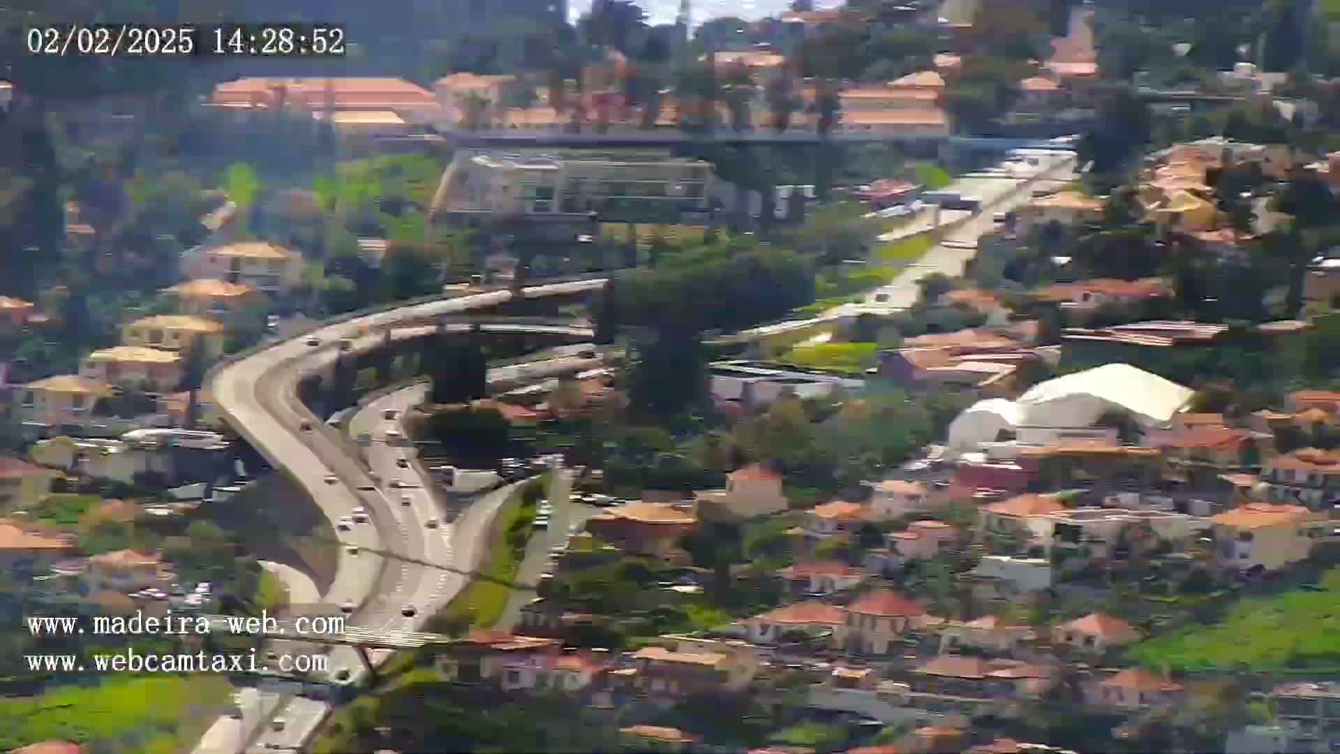 A winding highway with moderate traffic flows through a hillside neighborhood of closely packed, terracotta-roofed houses on a sunny day.